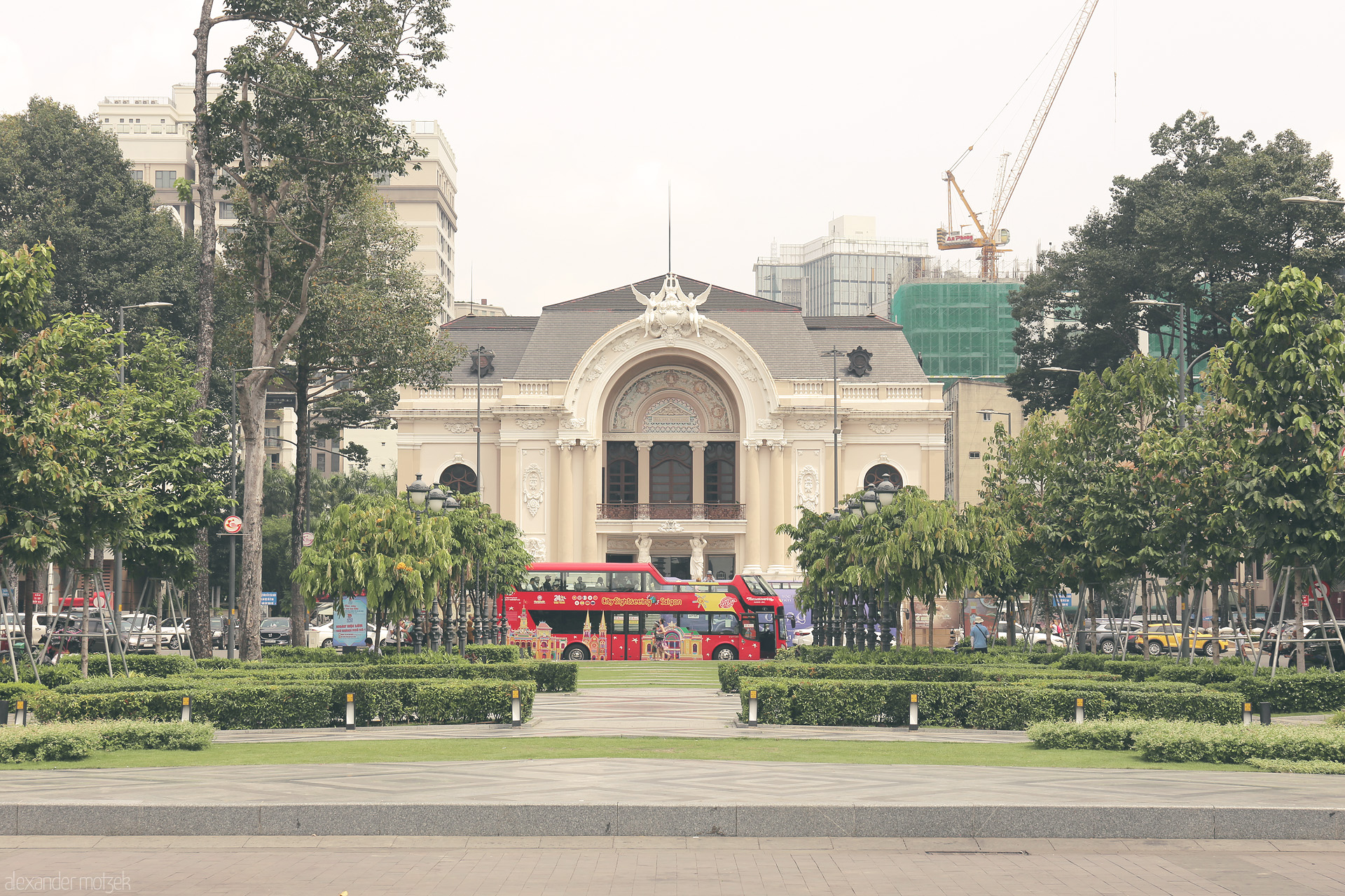 Foto von A pastel-hued Saigon Opera House stands behind lush trees and a red double-decker, blending old-world charm with modern city life in Ho Chi Minh City.