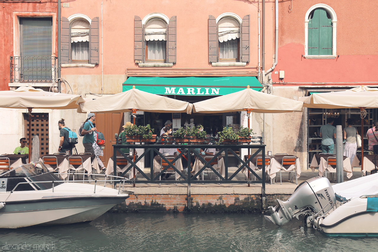 Marlin Venezia Vivente Foto von A quaint waterside café in Venice, with moored boats and bustling patrons adding to the serene Venetian charm.