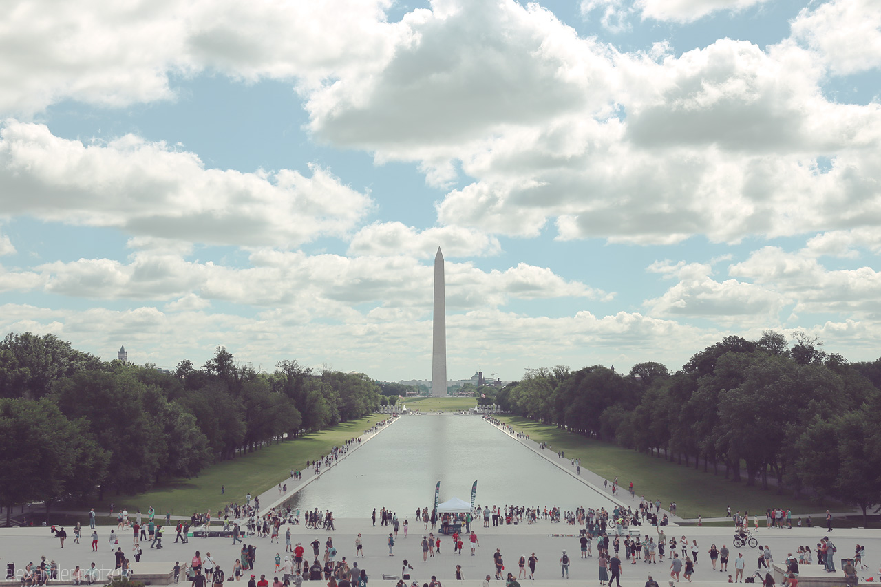 Monumental Stillness Foto von A serene morning view of the Washington Monument framed by lush greenery and a bustling crowd beneath a canopy of cotton clouds.