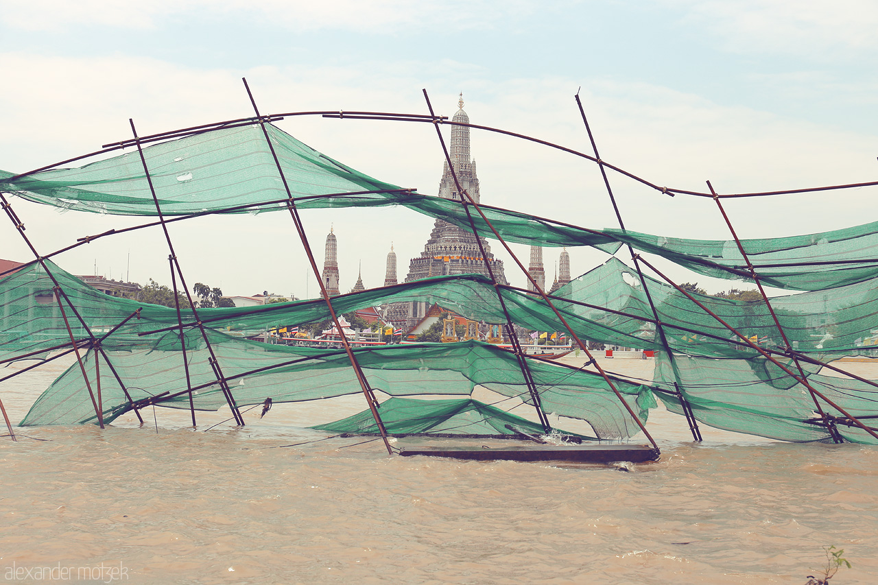 Veiled Reflections of Ratchawang Foto von A serene view of Phra Borom Maha Ratchawang across the river, framed by delicate green nets, capturing the essence of Bangkok's timeless beauty.