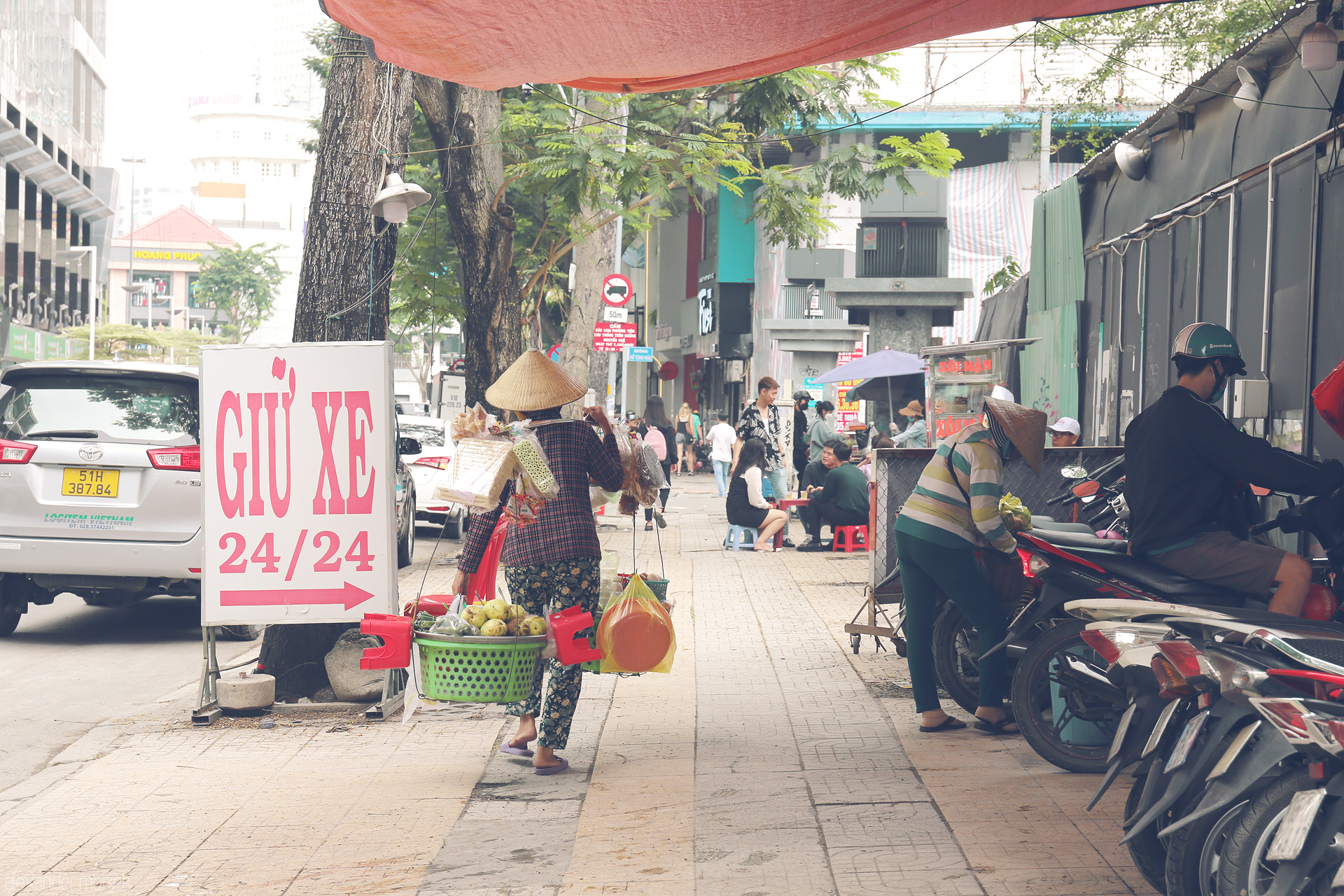 Saigon Sidewalk Symphony Foto von A street vendor in a nón lá weaves through Saigon’s bustling sidewalks, framed by local life, scooters, and a bold 'Giu Xe' sign.