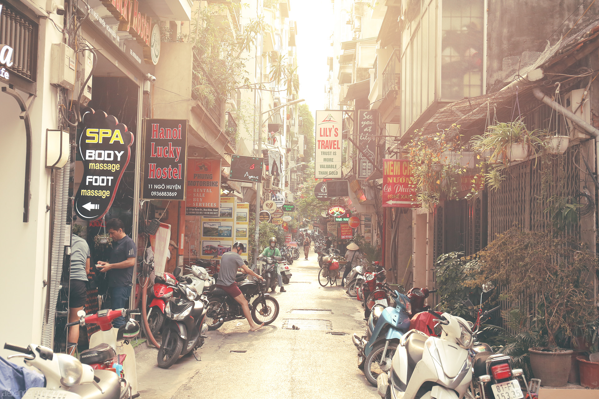 Foto von A sun-drenched alley in Phuong Hàng Trong, Hanoi, buzzing with scooters, neon signs, and local life blending tradition and travel.