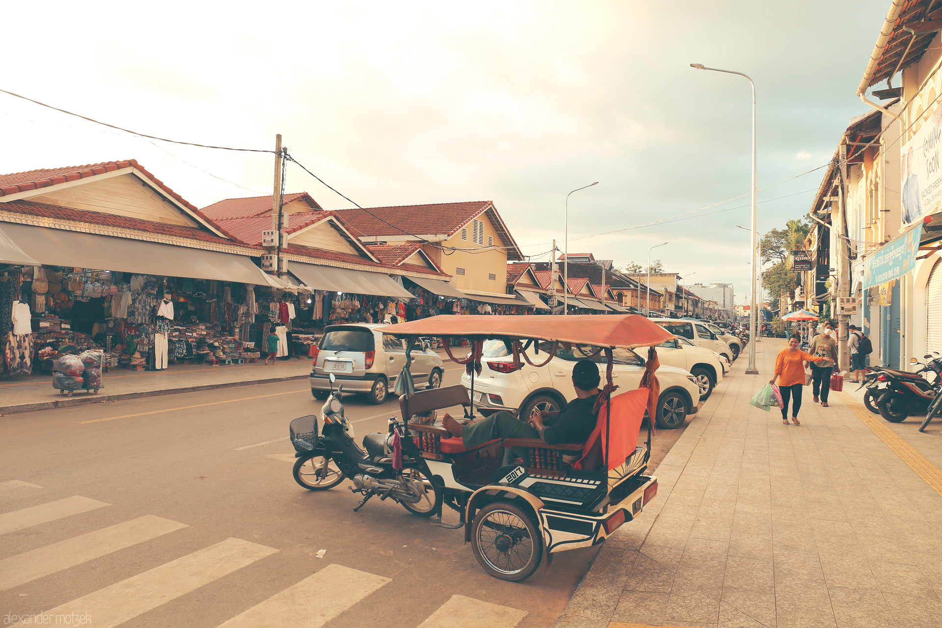 Tuk-Tuk Tranquility Foto von A tuk-tuk driver lounges by Siem Reap’s bustling Psar market, warm sunlight bathing local shops and vibrant street life.