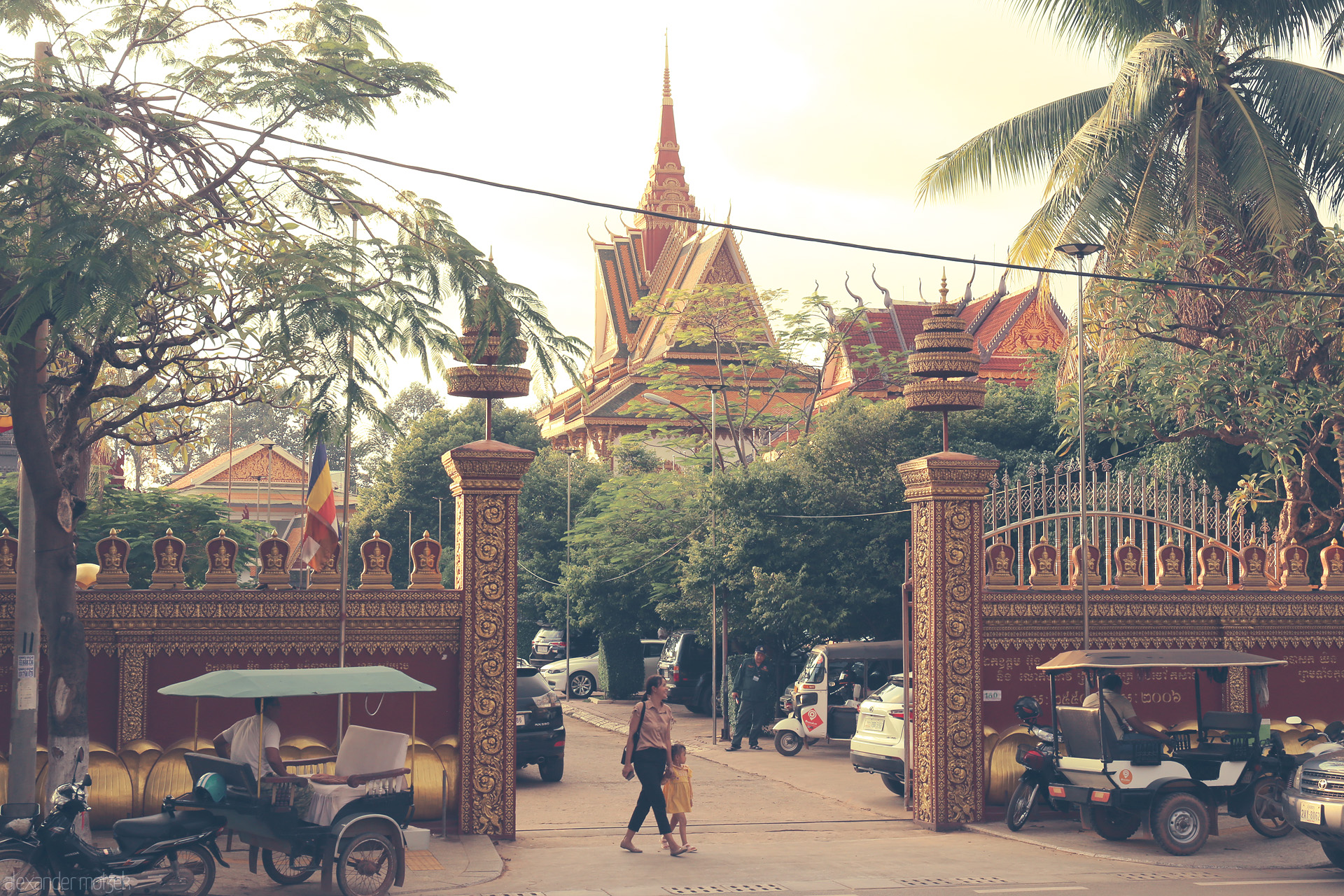 Angkor Twilight Gat Foto von A woman and child walk past golden gates and tuk-tuks, with a Khmer temple shimmering under Siem Reap’s lush palms and late sunlight.