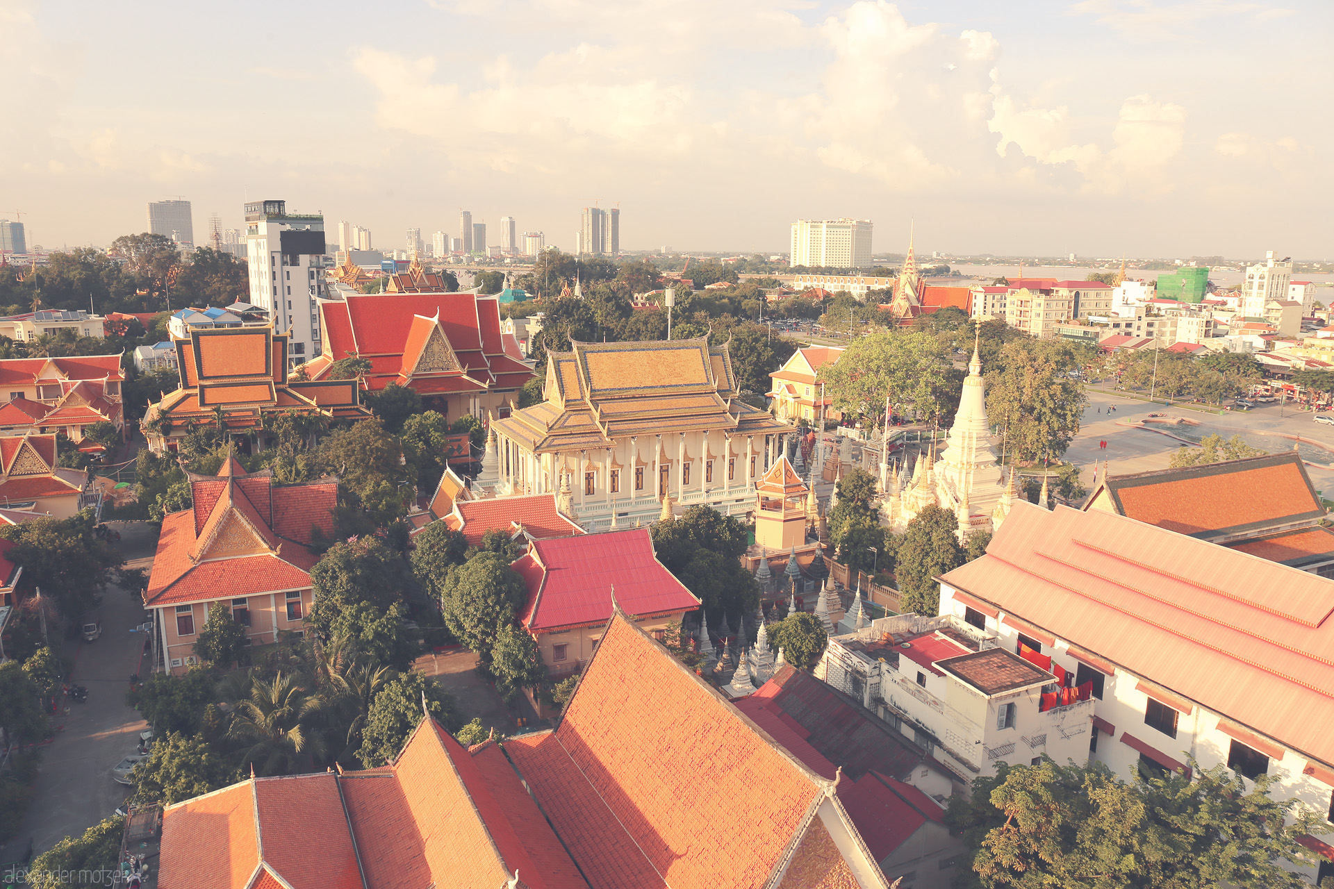 Pagodas in the Golden Light Foto von Aerial view of ornate temple roofs and stupas in Phnom Penh, Cambodia, basking in golden afternoon light against a modern city skyline.