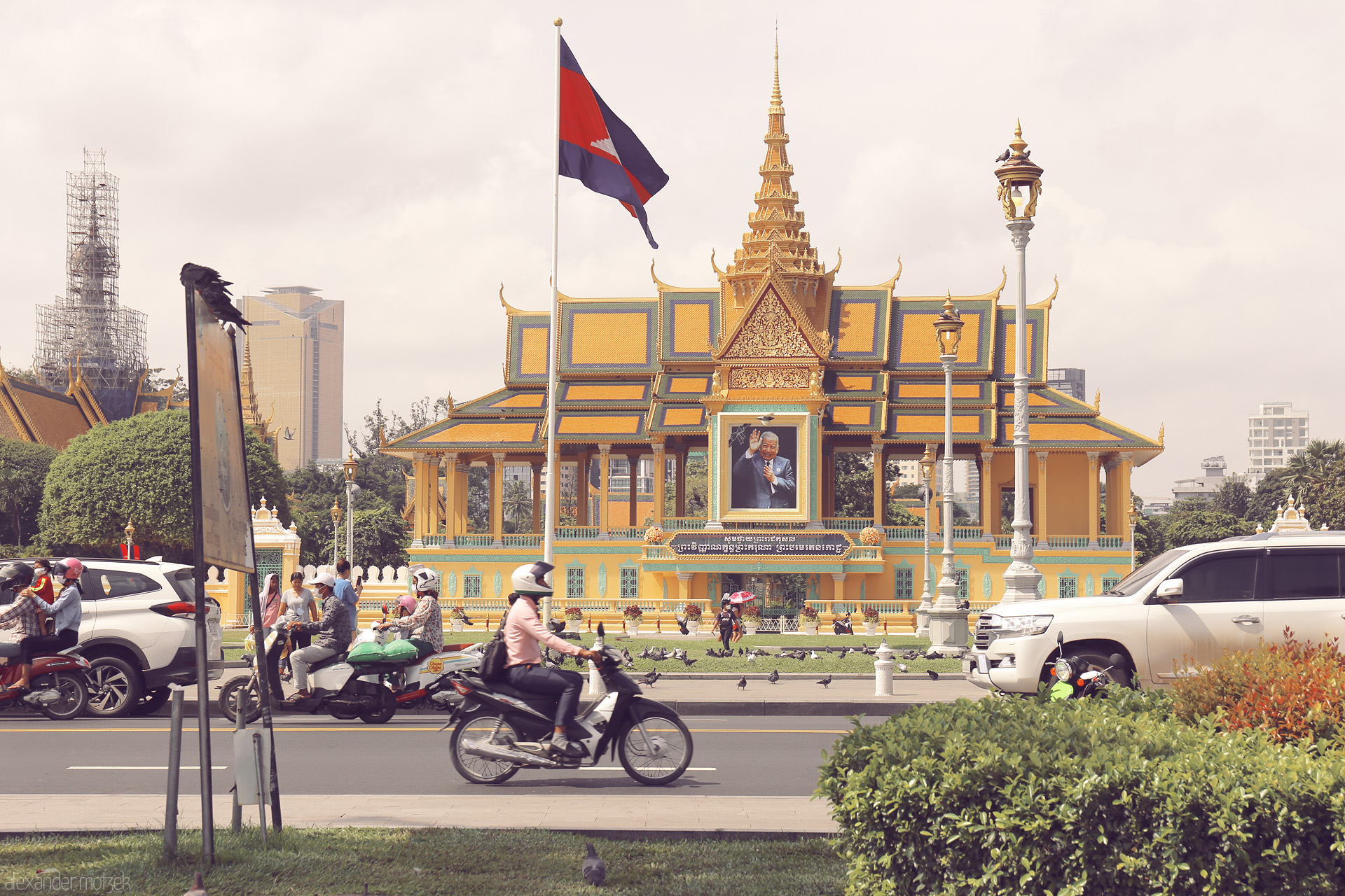 Motos & Monivong Foto von Beneath the royal gold of Phnom Penh, daily life streams by—scooters, locals, and pigeons beneath the Cambodian flag fluttering in the sun.