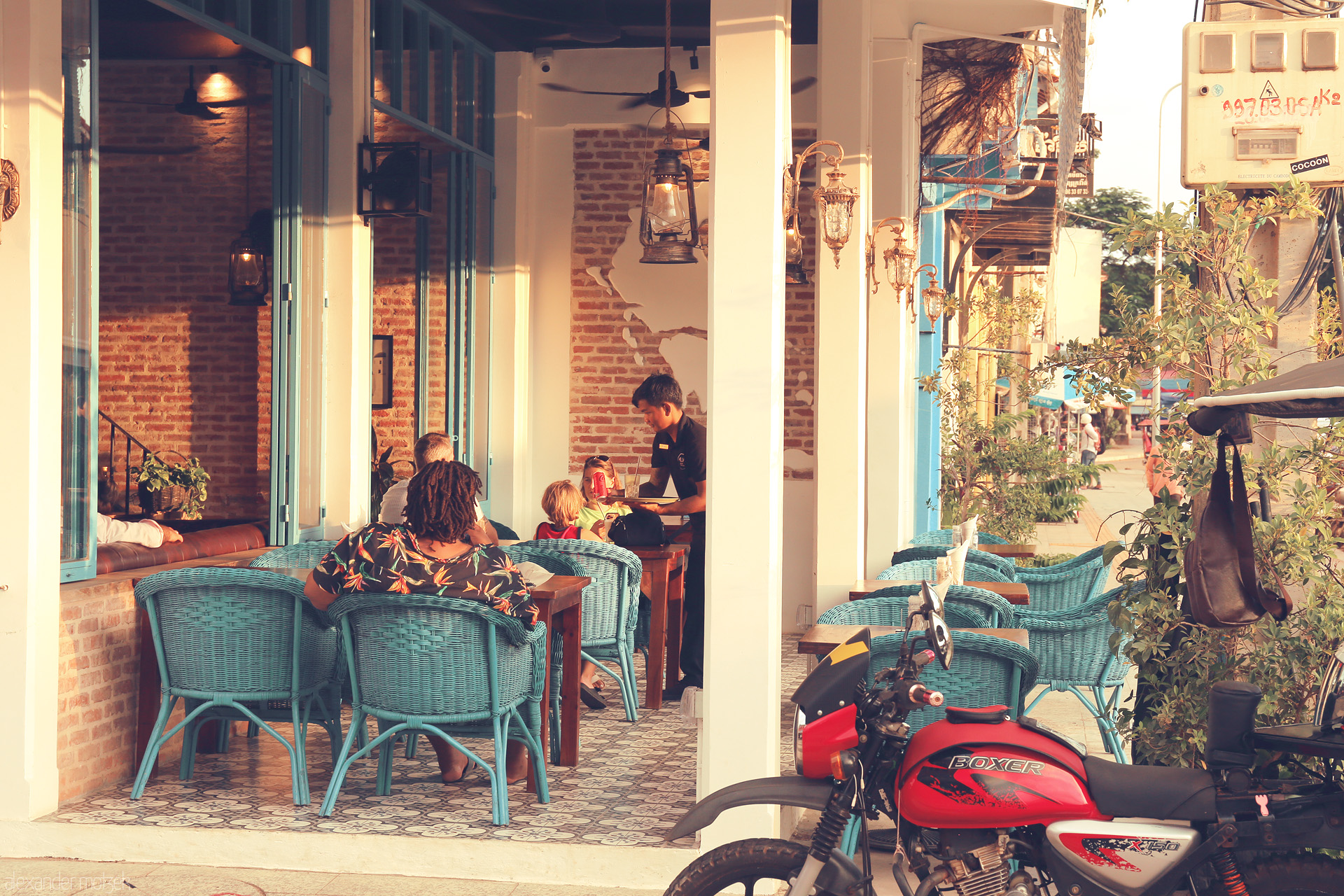 Angkor Apero Foto von Evening light spills onto a cozy Siem Reap café terrace, turquoise chairs, laughter, and a red motorbike capturing Khmer street charm.
