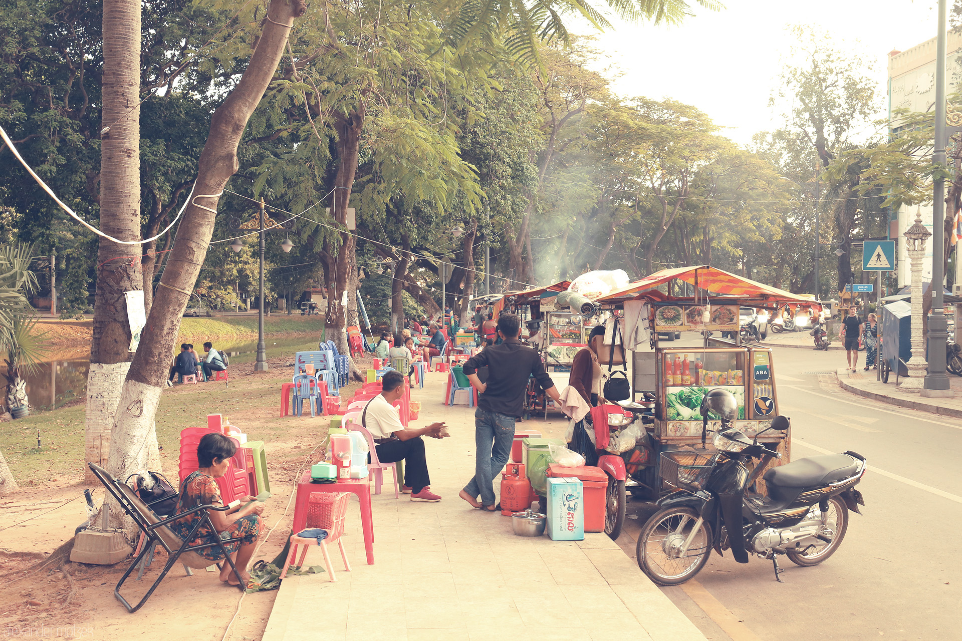 Chairs & Chai by the River Foto von Morning bustle in Siem Reap: street food carts, plastic chairs, and locals savor moments by the riverside under lush tropical trees.