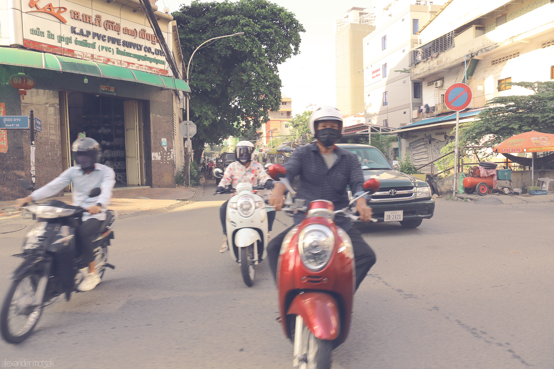 Rush on St. 390 Foto von Morning moto frenzy in Kandal: blurring scooters ride past shops and street signs on sunlit Street 390, Cambodia’s bustling daily rhythm.