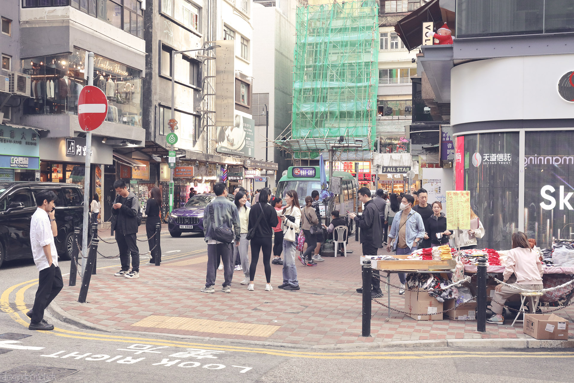 Foto von Crowded Wan Chai street corner, Hong Kong—sidewalk clothing stall, minibus stop, and green-scaffolded buildings in soft city haze.