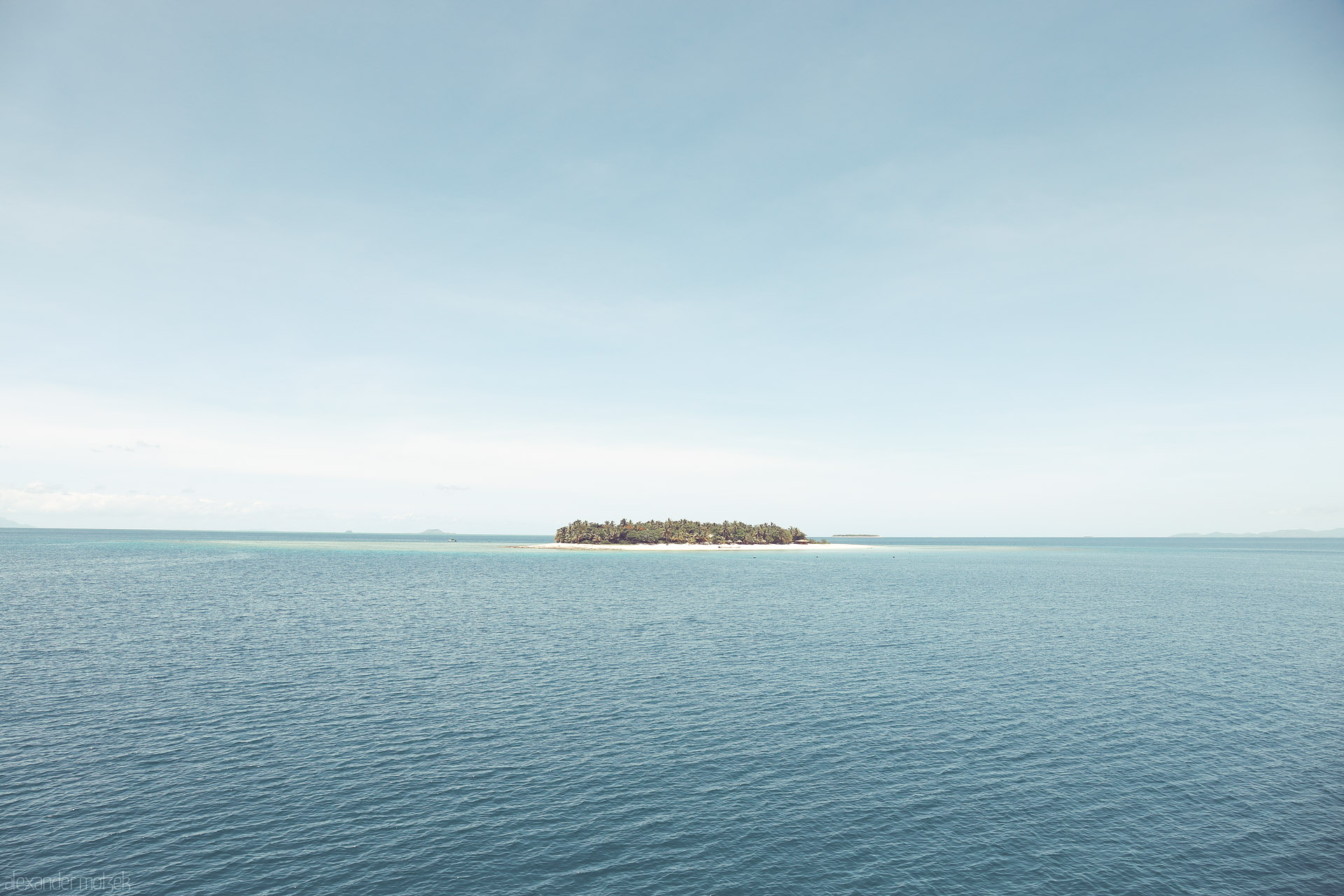 Foto von A lone palm-crowned motu floats on glassy Pacific blues beneath a pale sky—Mamanuca Islands, Fiji.