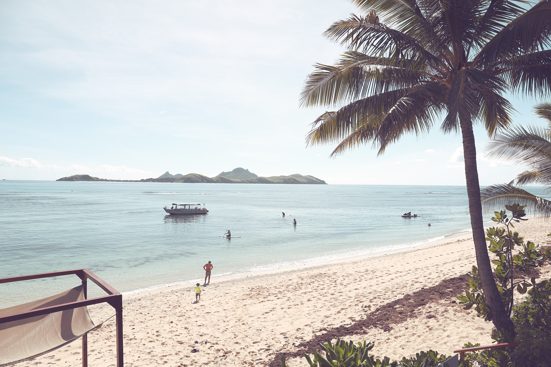 Foto von Tokoriki, Fiji—palm-framed beach, glassy lagoon with a boat, paddleboarders and swimmers, Mamanuca isles beyond.