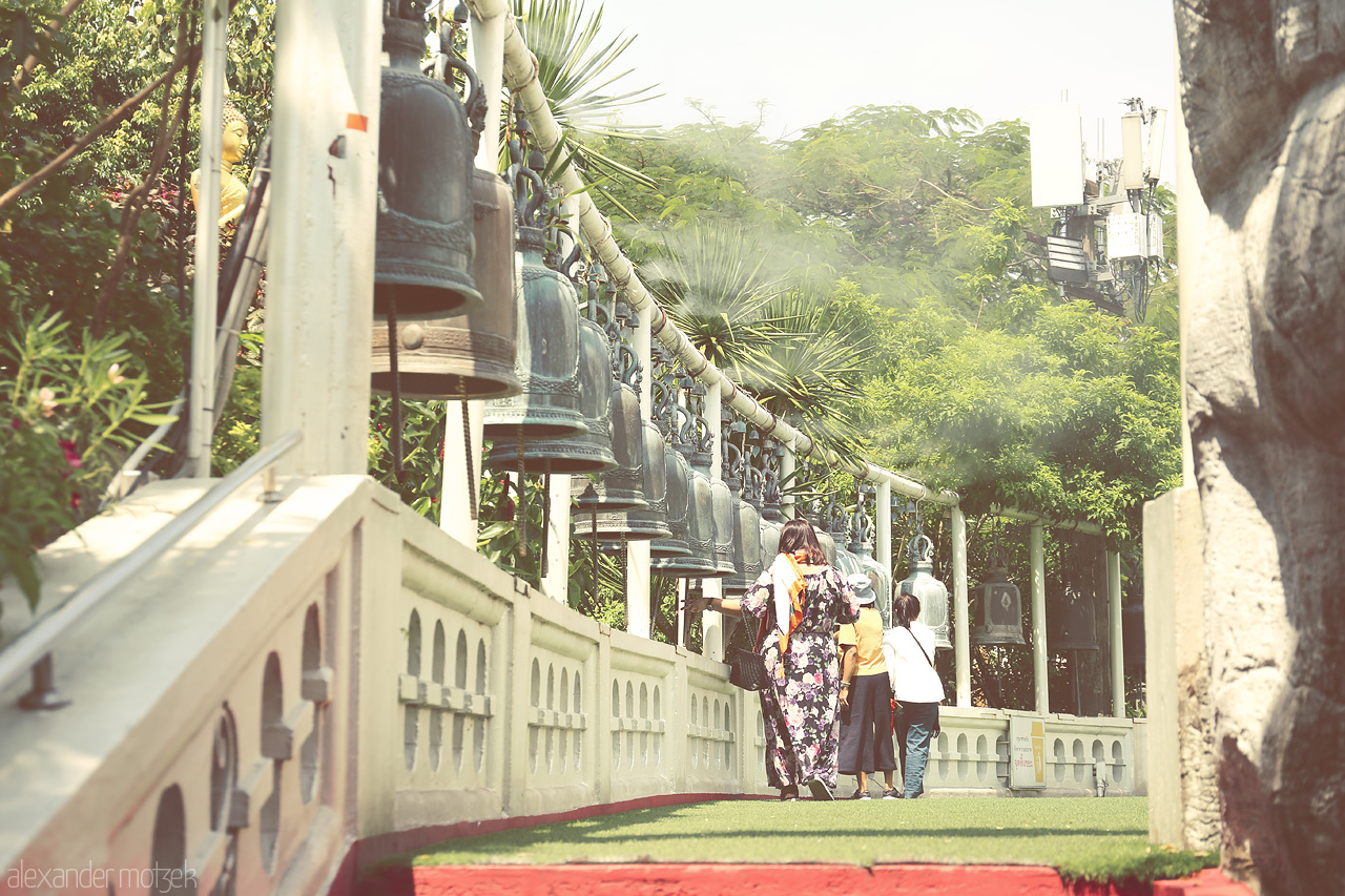 Echoes of Ban Bat Foto von In Ban Bat, Bangkok, visitors stroll by a tranquil row of bells, capturing a serene mix of tradition and nature.