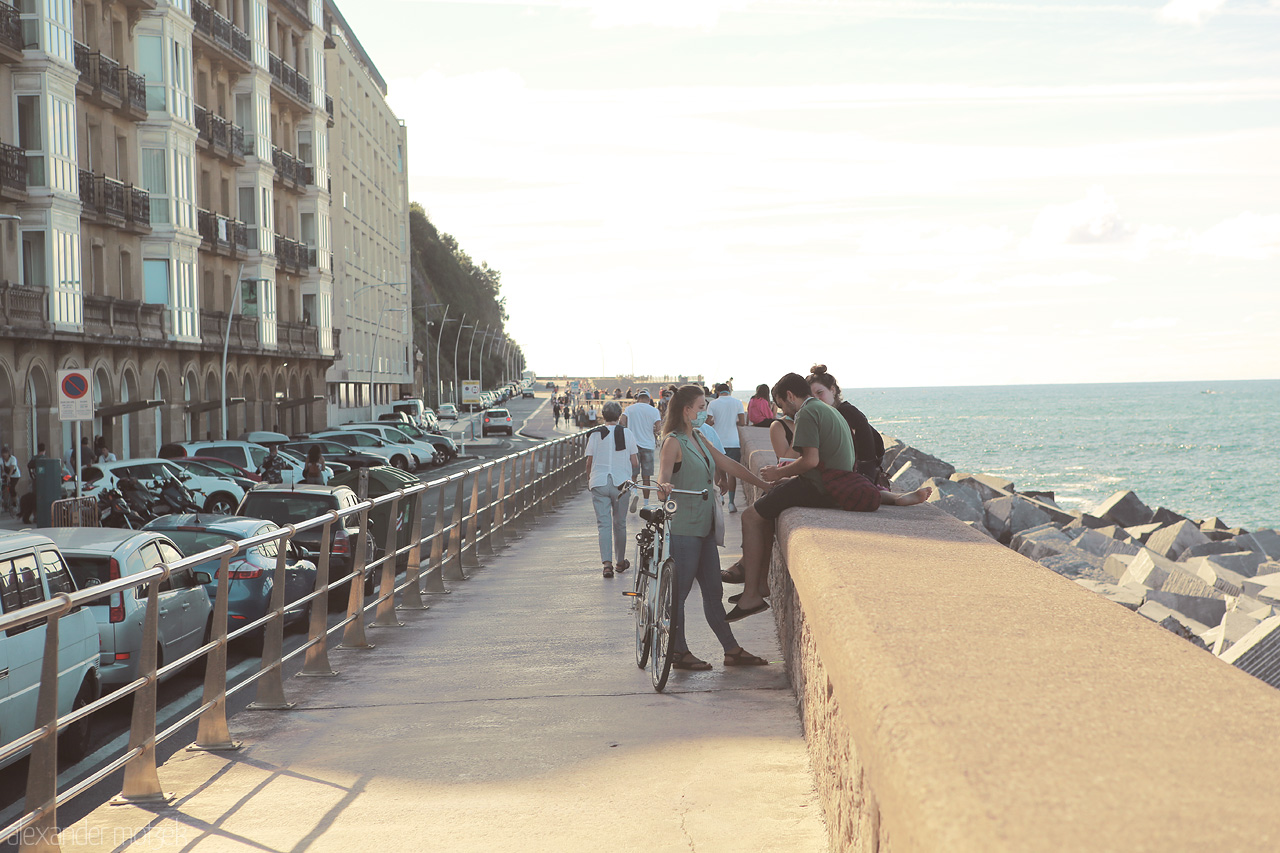 Donostia Dusk Stroll Foto von Pedestrians enjoying a seaside promenade in San Sebastián, with a backdrop of the ocean and lined cars.
