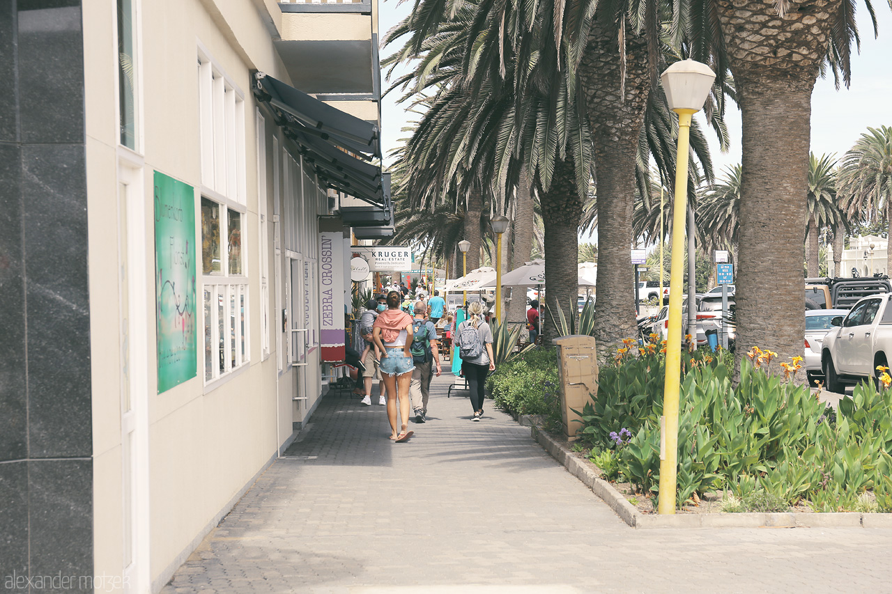 Swakopmund Strolls Foto von Pedestrians navigate a palm-lined boulevard, capturing the quintessence of Swakopmund.