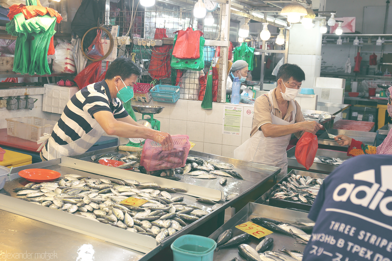 Pasar Ikan Hustle Foto von At a bustling Singapore market, vendors prepare fresh fish, surrounded by colorful bags and produce. A snapshot of local daily life.