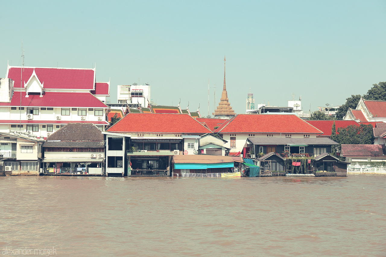 Chao Phraya Serenity Foto von Tranquil views of vibrant red-roofed homes along the Chao Phraya River in Khlong San, Bangkok, with a distant temple spire.