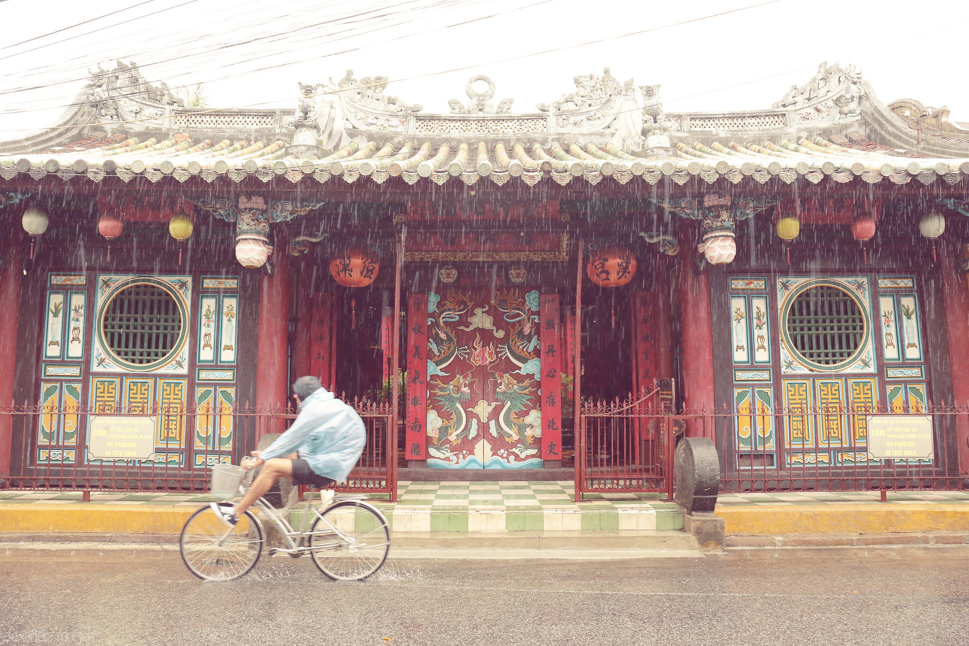 Lanterns in the Storm Foto von A cyclist glides past the dragon doors of a Hoi An temple, rain dancing off ancient lanterns in Vietnam’s poetic embrace.