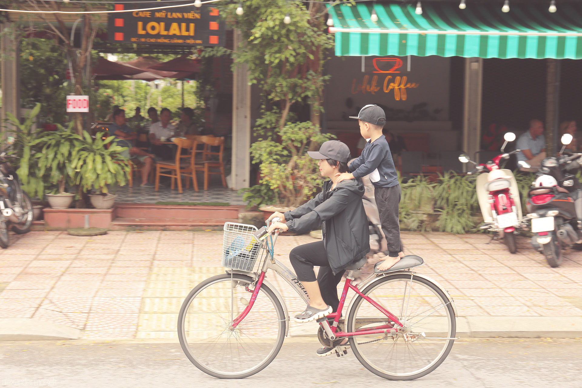 Cafe Cruise Foto von A mother and child glide by a cozy Hoi An café, balancing daily life and joy on a bicycle in Vietnam’s lantern-lit town.