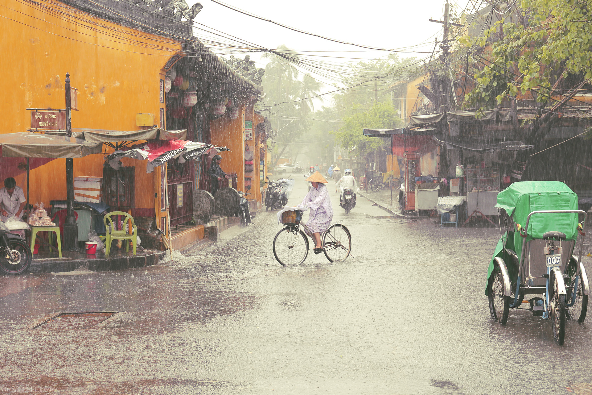 Conical Hat Serenade Foto von A solitary cyclist glides through Hoi An in the rain, framed by yellow walls, lanterns, and daily life under monsoon skies.