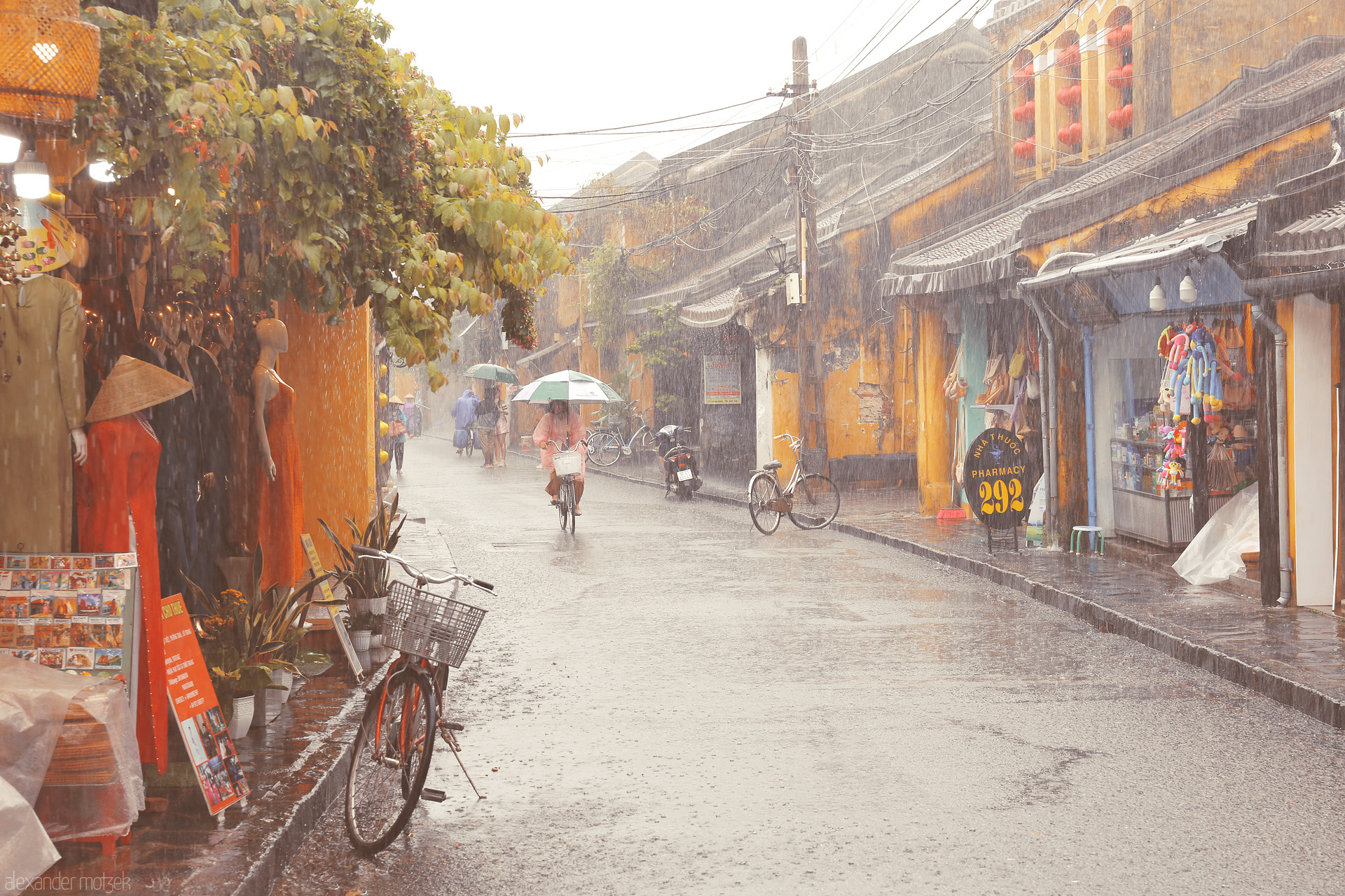 Áo Dài & Umbrellas Foto von Cyclists and street life soak in golden rain showers in Hoi An, Vietnam, blending áo dài elegance and vibrant lantern-lit shops.