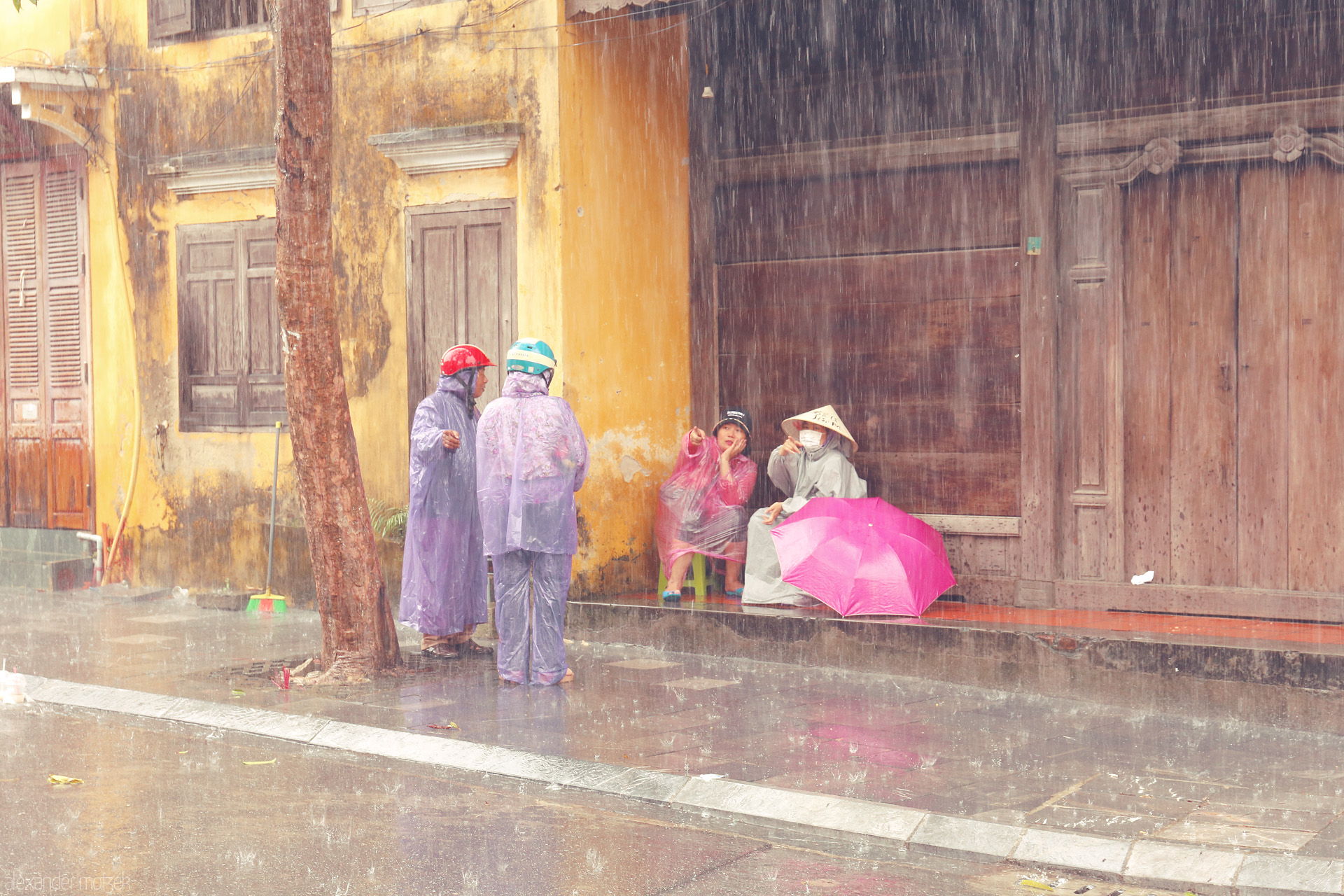 Mua Hoi An Whispers Foto von Locals pause under the rain in Hoi An, Vietnam, bright raincoats and a pink umbrella glowing against ancient yellow walls.