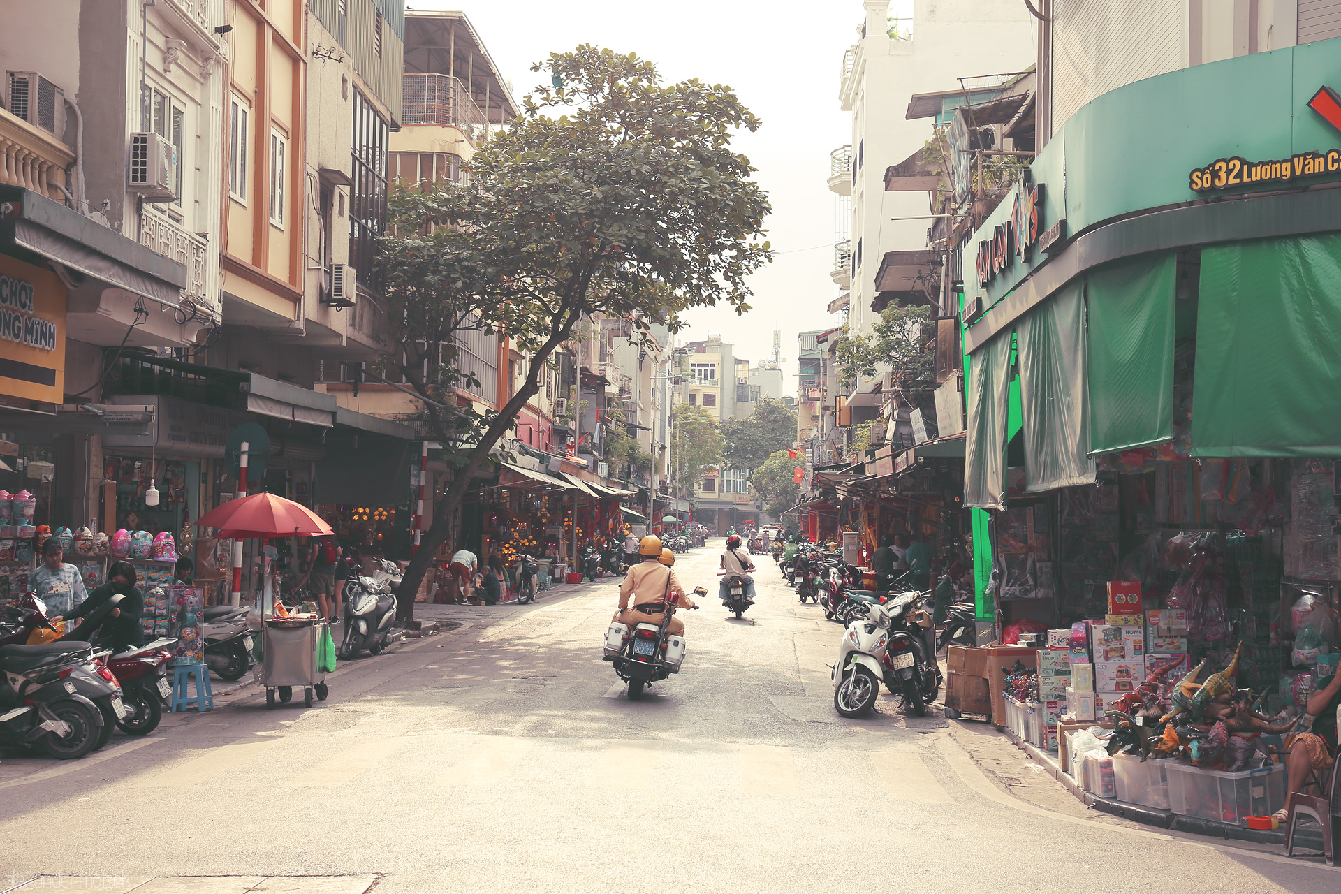 Scooters & Shadows Foto von Morning scooters glide through sun-drenched streets of Phuong Hàng Dào, Hanoi, where market life hums beneath leafy branches.
