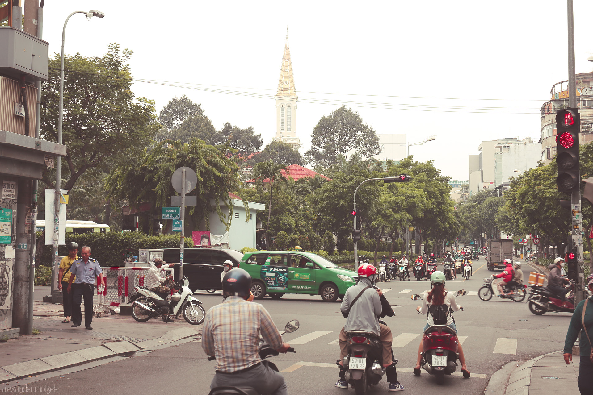 Scooters & Spires Foto von Motorbikes pause at a red light on Pham Ngu Lão, Saigon, as a church spire rises above lush trees in Ho Chi Minh City.