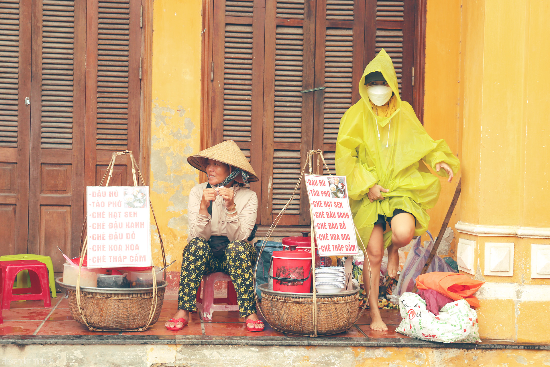 Chè & Golden Walls Foto von Streetside chè sellers pause on a rainy day, yellow ancient walls of Hoi An framing everyday life and local flavors.